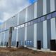 Außenansicht eines Logistikzentrums im Bau mit vertikal gestalteter Metallfassade und Fensterband unter wolkigem Himmel