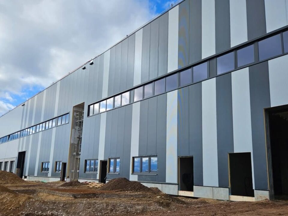 Außenansicht eines Logistikzentrums im Bau mit vertikal gestalteter Metallfassade und Fensterband unter wolkigem Himmel